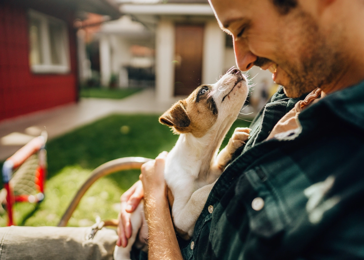 Man smiling at a puppy on his lap in a sunny backyard.