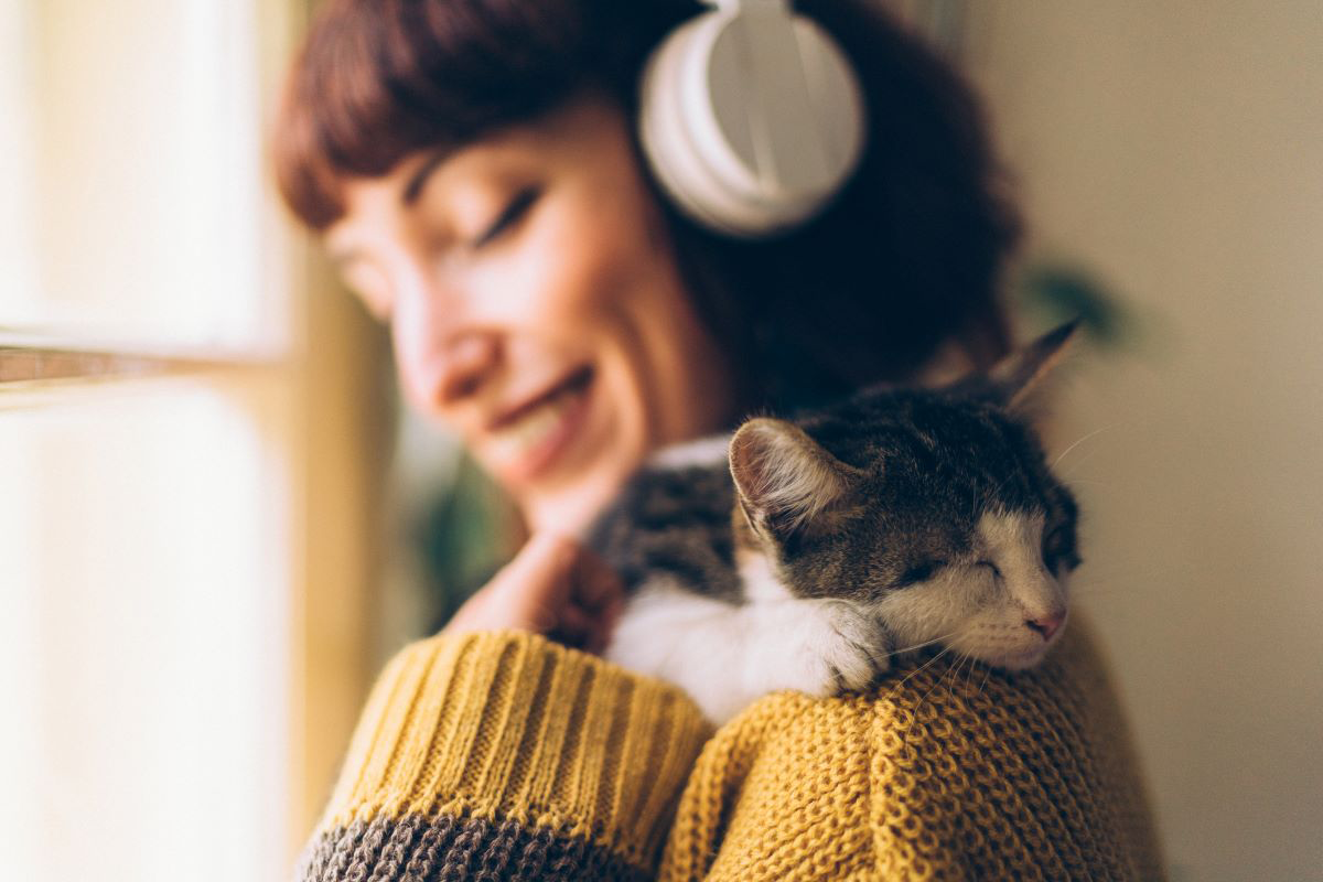 Smiling person wearing headphones with a sleeping cat on their shoulder.