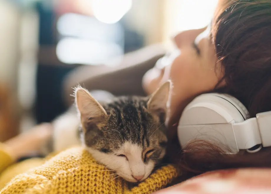Woman lying down with headphones, cat sleeping on her chest.