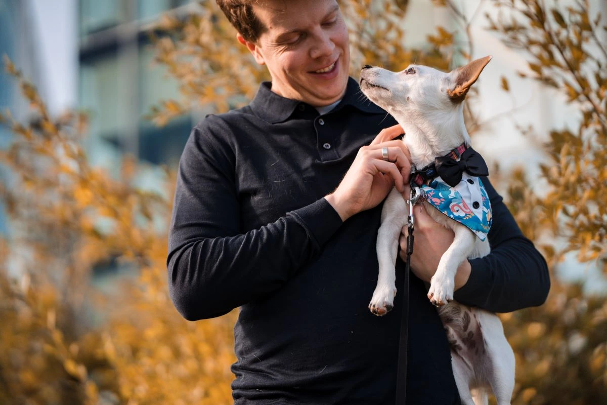 Man in black shirt holding a small dog in a tuxedo bandana.