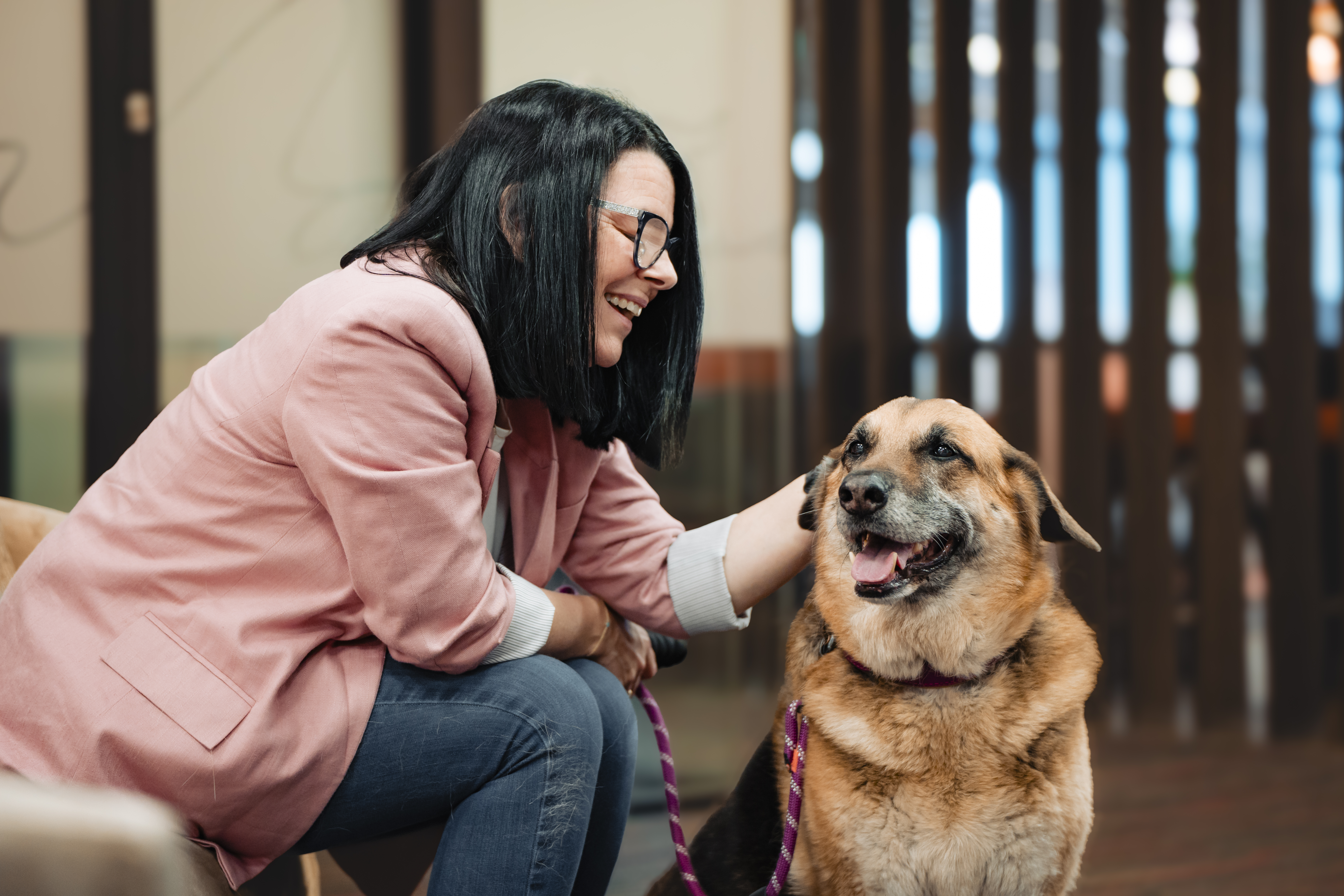 Mulher de blazer rosa sorrindo para um cachorro dentro de casa.